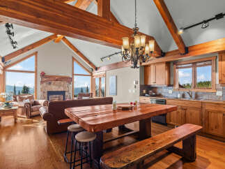 Experience breathtaking mountain views from this stunning living and dining area, featuring a grand stone fireplace and vaulted wooden beam ceilings.