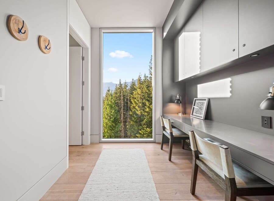 Experience breathtaking views from this modern home office, featuring a large picture window overlooking a lush forest and distant mountains.