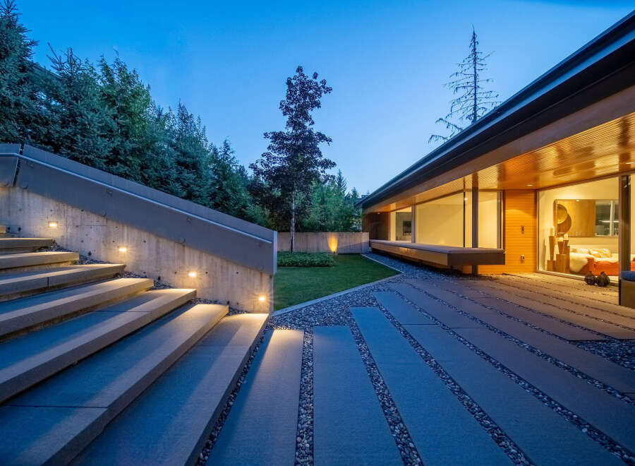Experience modern luxury with this stunning property featuring a sleek concrete staircase illuminated by subtle lighting, leading to a contemporary home with expansive glass windows.