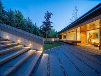 Experience modern luxury with this stunning property featuring a sleek concrete staircase illuminated by subtle lighting, leading to a contemporary home with expansive glass windows.