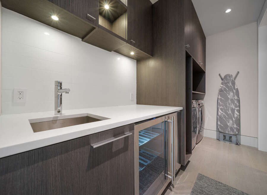 This modern laundry room features a sleek white countertop with an undermount sink and a chrome faucet, complemented by dark wood cabinetry.