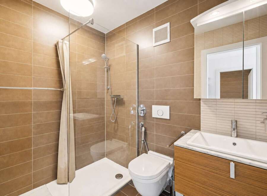 This modern bathroom features a sleek design with warm brown tiled walls and a spacious walk-in shower enclosed by a glass panel and a light-colored curtain.
