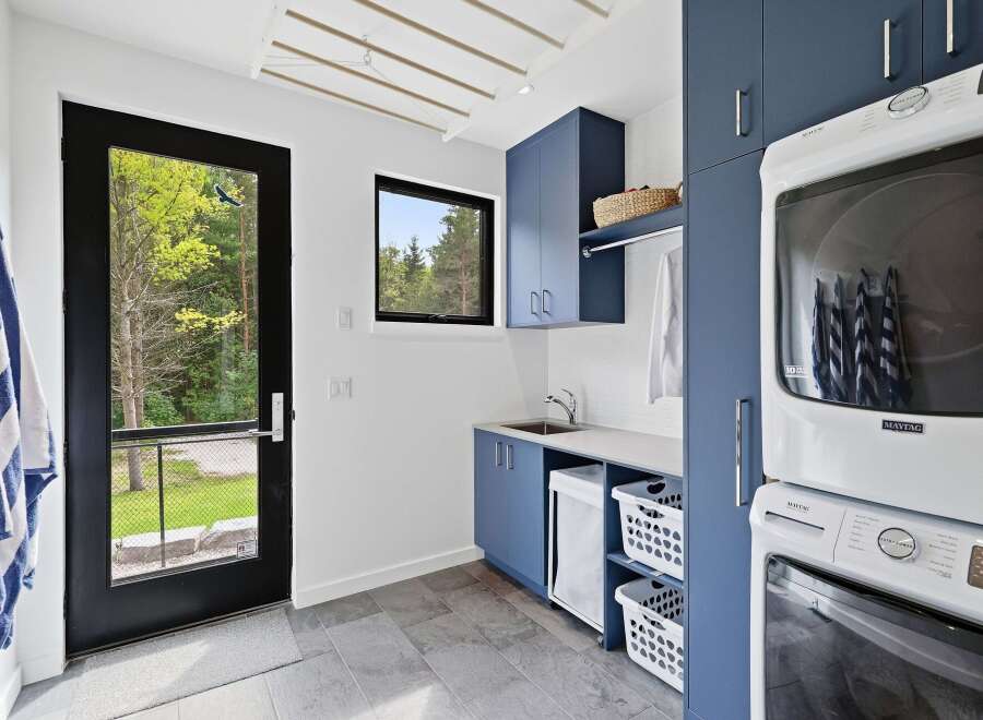 This modern laundry room features sleek navy blue cabinetry, a stainless steel sink, and a stacked Maytag washer and dryer.