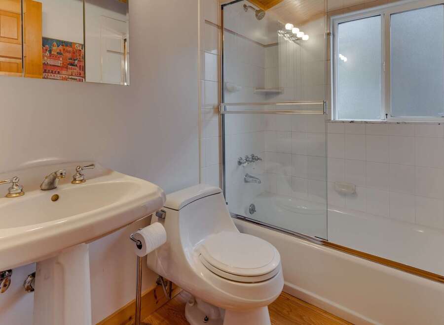 This charming bathroom features a pedestal sink with elegant gold-accented fixtures, a modern toilet, and a pristine white bathtub with a glass shower enclosure.