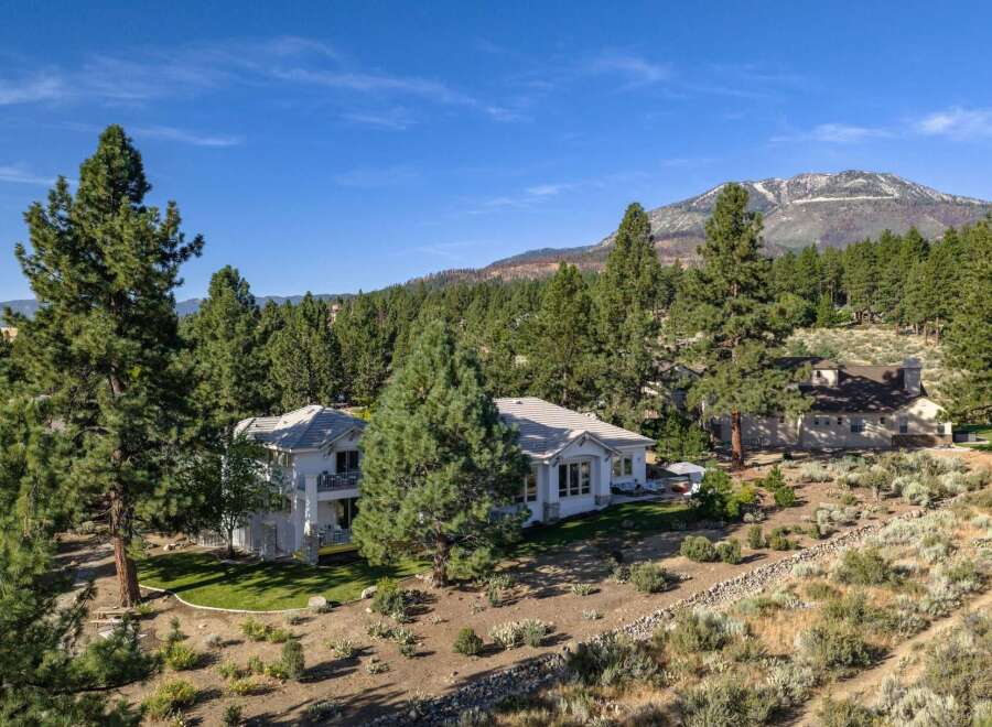 Nestled amidst towering pine trees and offering breathtaking mountain views, this stunning property boasts a modern white home with multiple balconies and a lush green lawn.