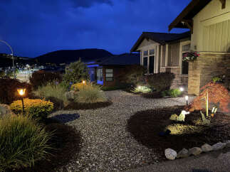 Experience the charm of this beautifully illuminated home at dusk, featuring a welcoming pathway lined with decorative gravel and lush landscaping.
