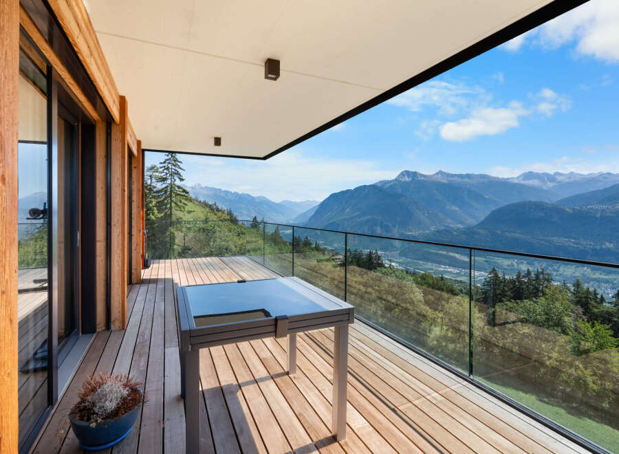 Experience breathtaking mountain vistas from this stunning modern balcony, featuring a sleek glass railing and a stylish outdoor table.