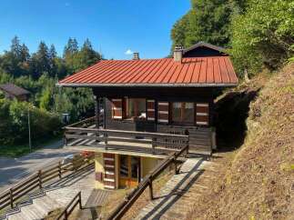 Charming wooden chalet nestled on a hillside, offering a rustic retreat with a vibrant red roof and a spacious wooden deck perfect for enjoying the surrounding nature.