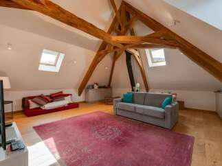 Discover this charming attic living space, featuring exposed wooden beams and a cozy seating area with a vibrant red floor cushion and a comfortable grey sofa.