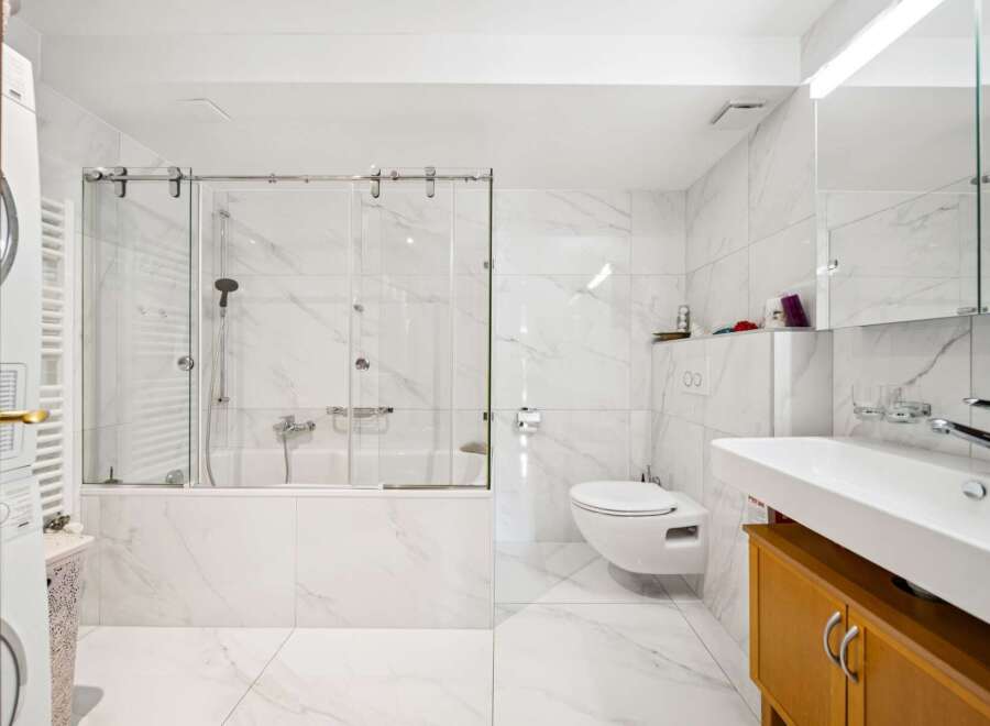 This modern bathroom features a sleek bathtub with a glass enclosure, a wall-mounted toilet, and a spacious vanity with a large sink.