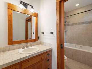 This bathroom features a warm wooden vanity with a tiled countertop and a white undermount sink, complemented by a rustic wooden-framed mirror.