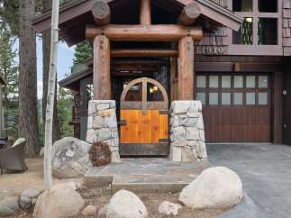 Experience rustic charm with this stunning log home featuring a unique arched wooden gate entrance, complemented by natural stone pillars and a welcoming wreath.