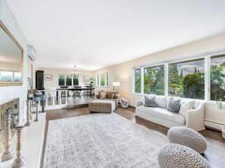 This spacious open-concept living and dining area boasts abundant natural light streaming through large windows, offering picturesque views of lush greenery.