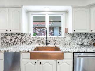 This kitchen showcases a stunning copper farmhouse sink, complemented by elegant white cabinetry and a dramatic granite backsplash.