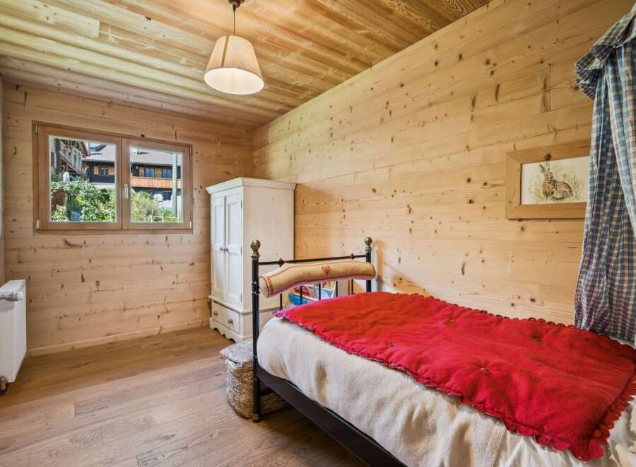 Experience rustic charm in this cozy bedroom, featuring warm wooden walls and ceiling, a comfortable bed with a vibrant red throw, and a charming window overlooking the picturesque surroundings.