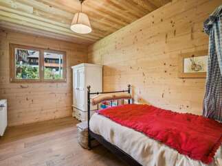Experience rustic charm in this cozy bedroom, featuring warm wooden walls and ceiling, a comfortable bed with a vibrant red throw, and a charming window overlooking the picturesque surroundings.