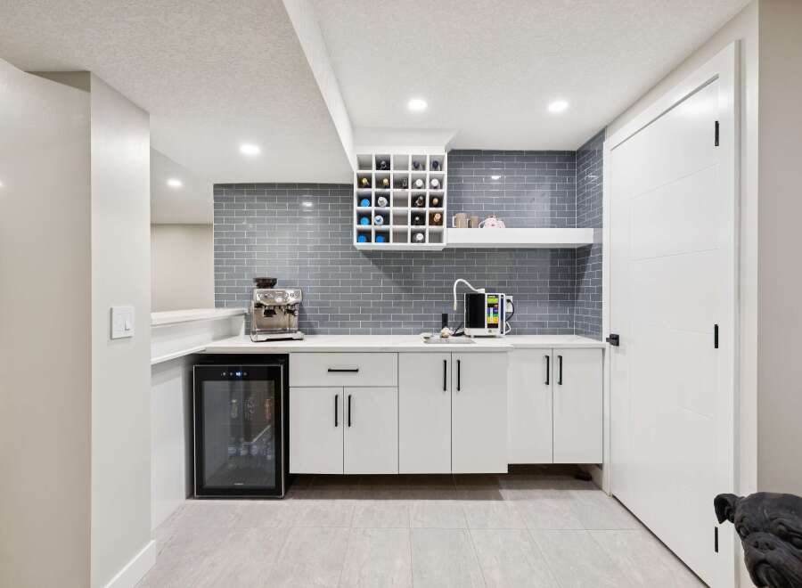 This modern basement bar features a sleek white countertop with a grey subway tile backsplash, complemented by a built-in wine rack and a compact beverage refrigerator.
