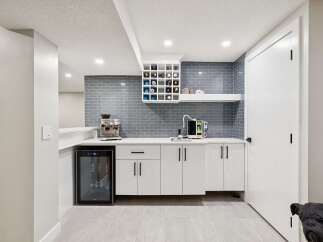 This modern basement bar features a sleek white countertop with a grey subway tile backsplash, complemented by a built-in wine rack and a compact beverage refrigerator.