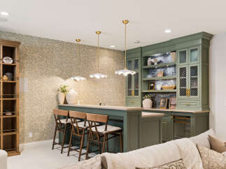 This inviting basement bar area features a stylish green cabinetry setup with glass-fronted cabinets and open shelving, complemented by a sleek countertop and comfortable bar stools.