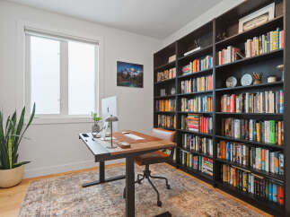 This home office features a modern standing desk with a computer setup, complemented by a rich leather chair and a large, dark wood bookshelf filled with a diverse collection of books.
