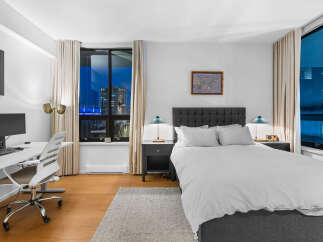 Experience modern city living in this stylish bedroom featuring a comfortable bed, a dedicated workspace with a computer, and large windows offering stunning night views.