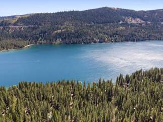 Experience breathtaking lakefront living with this stunning aerial view of a serene blue lake surrounded by lush green pine forests.