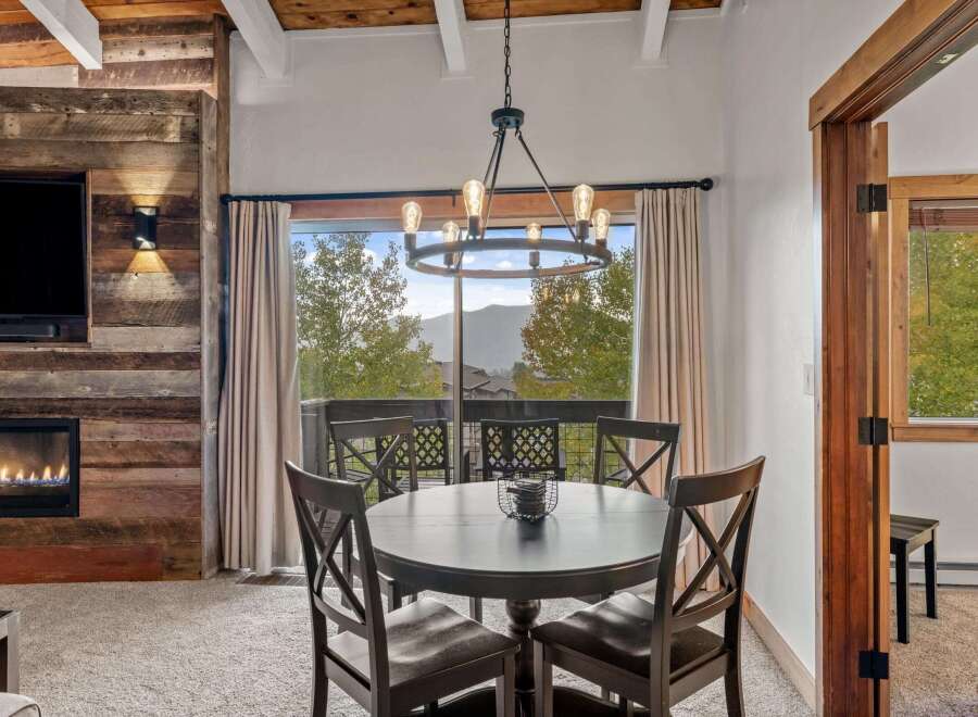 Experience cozy mountain living in this charming dining area, featuring a rustic wood-paneled accent wall with a modern fireplace and a large window offering breathtaking views of the surrounding landscape.