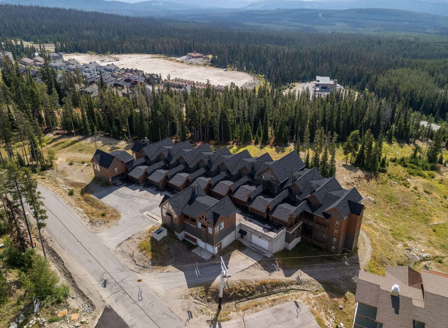 Discover this stunning mountain property featuring multiple townhome-style residences with dark pitched roofs and warm wood exteriors, nestled amidst a dense evergreen forest.