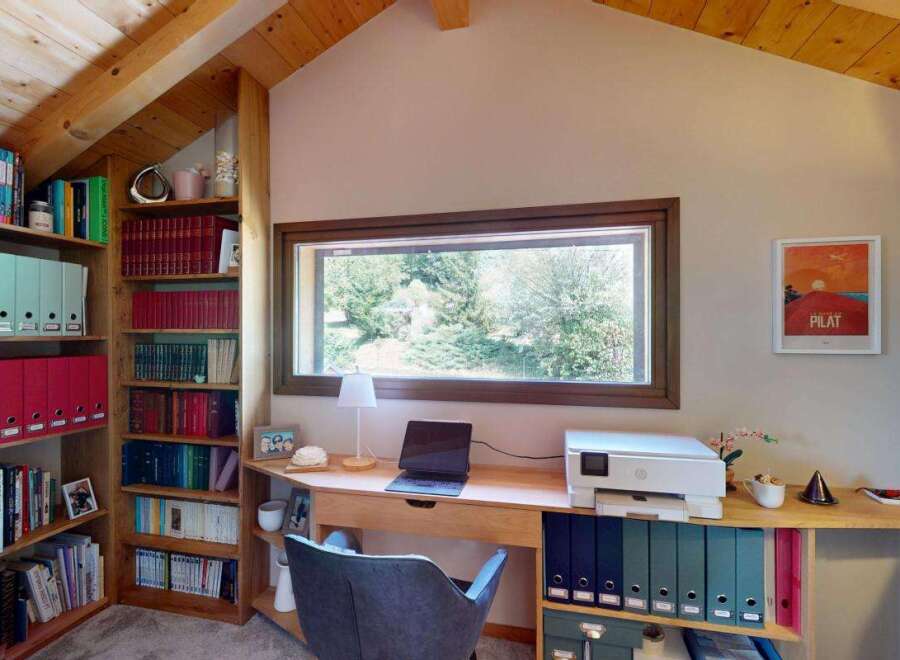 Discover a cozy home office space featuring a large window with scenic views, a built-in bookshelf filled with binders and books, and a functional desk area with a laptop and printer.