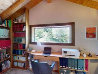 Discover a cozy home office space featuring a large window with scenic views, a built-in bookshelf filled with binders and books, and a functional desk area with a laptop and printer.
