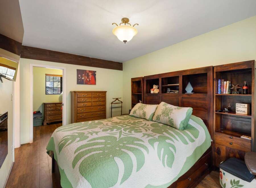 This charming bedroom features a spacious bed with a tropical-themed quilt and matching pillows, complemented by a large wooden dresser and built-in bookshelves.
