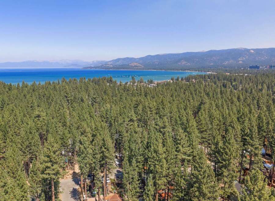 Experience the ultimate lakeside living with this stunning aerial view of a property nestled amidst a dense pine forest, offering breathtaking vistas of a vast blue lake and distant mountains.