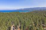 Experience the ultimate lakeside living with this stunning aerial view of a property nestled amidst a dense pine forest, offering breathtaking vistas of a vast blue lake and distant mountains.