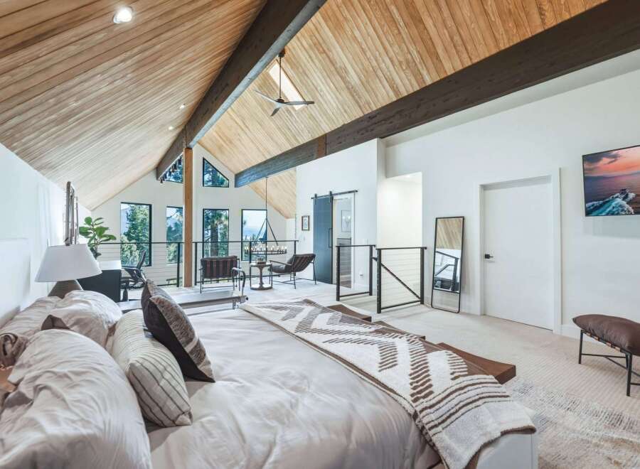 Experience luxurious mountain living in this stunning master suite, featuring a vaulted wood-paneled ceiling with exposed beams and expansive windows offering breathtaking forest views.
