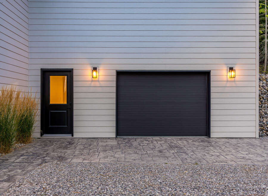 This modern home features a sleek dark garage door and a welcoming black entry door with a glass pane, illuminated by stylish outdoor lighting.