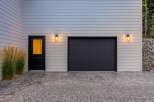 This modern home features a sleek dark garage door and a welcoming black entry door with a glass pane, illuminated by stylish outdoor lighting.