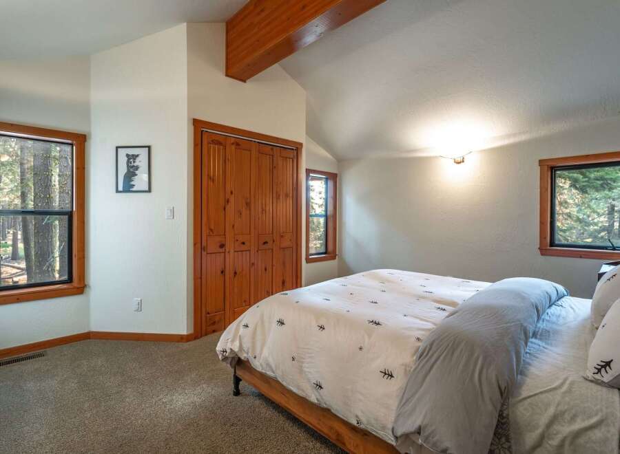 Experience cozy cabin living in this charming bedroom featuring a vaulted ceiling with exposed wooden beams and ample natural light from multiple windows.