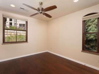 This room features a modern ceiling fan and two large windows, one offering views of a building and the other of lush pine trees.