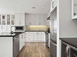 This modern kitchen features sleek white cabinetry, a spacious island with a granite countertop, and stainless steel appliances, including a built-in microwave and a French door refrigerator.