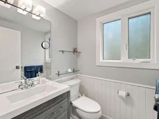 This modern bathroom features a sleek vanity with a white countertop and gray wood-grain cabinets, complemented by a large mirror and stylish light fixtures.