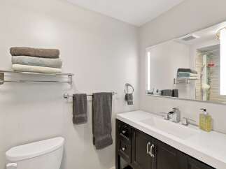 This modern bathroom features a sleek vanity with a white countertop and dark wood cabinets, complemented by a large illuminated mirror.