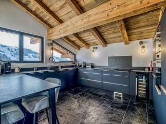 Experience luxury mountain living in this stunning kitchen featuring a dramatic vaulted ceiling with exposed wooden beams and a breathtaking panoramic view of snow-capped mountains through a large window.