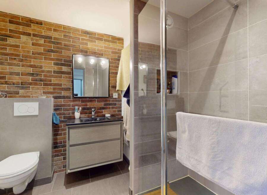 This modern bathroom features a striking exposed brick accent wall, complemented by sleek grey tiles and a floating vanity.