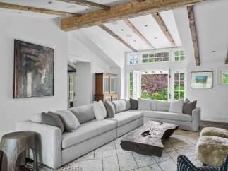 Experience rustic elegance in this spacious living room, featuring exposed wooden beams and a vaulted ceiling that enhance the natural light pouring in from the large windows.