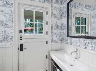 Experience rustic charm with this beautifully appointed bathroom featuring a unique Dutch door and striking toile wallpaper depicting a serene mountain landscape.
