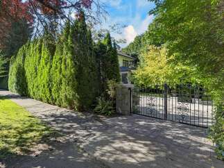 Discover this exquisite property, offering a grand entrance through an ornate black metal gate, flanked by a meticulously manicured hedge and mature trees.