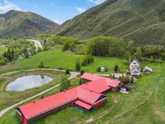 Discover this expansive ranch property nestled in a picturesque valley, featuring a large red-roofed barn, a charming main house with a wraparound deck, and a serene pond.