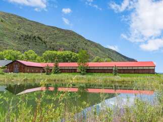 Experience serene living with this stunning property featuring a long, rustic barn with a vibrant red roof, nestled beside a tranquil pond reflecting the lush green landscape and a majestic mountain backdrop.