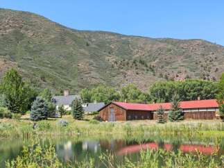 Nestled against a picturesque mountain backdrop, this property features a charming farmhouse and a rustic barn with a vibrant red roof, all overlooking a serene pond.