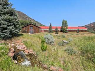 Discover this expansive property featuring a charming rustic barn with a bright red roof, set against a backdrop of rolling hills and clear blue skies.